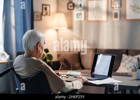 Rear view of senior man using wheelchair and working online on laptop at the table in the room Stock Photo