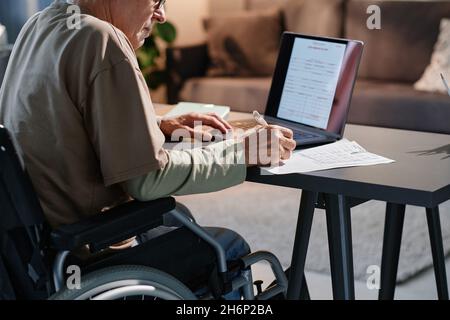 Senior man making notes in documents while sitting at the table working online on laptop in the room Stock Photo