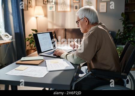 Senior man in eyeglasses using wheelchair sitting at the table and typing documents online on laptop, he working in the room Stock Photo