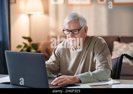 Senior man in eyeglasses using laptop for online work while sitting at the table in the room at home Stock Photo