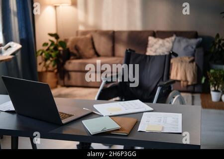 Image of workplace with laptop on it and wheelchair near the table in empty living room Stock Photo