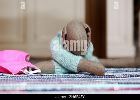 Kuscheltier süßer Hase mit langen Ohren und hellblauem Wollpulli, liegend auf einem bunten Teppich. Daneben eine rosa farbene Maske (Mundschutz). Stock Photo