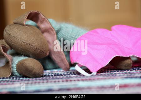 Kuscheltier süßer Hase mit langen Ohren und hellblauem Wollpulli, liegend auf einem bunten Teppich. Daneben eine rosa farbene Maske (Mundschutz). Stock Photo