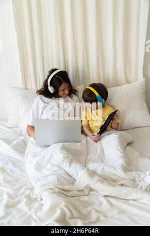 Mom and Son work and study lying together in bed using laptop computer and digital tablet in bedroom Stock Photo
