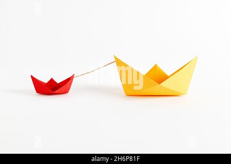Origami big yellow paper boat pulls the small red boat with rope. Support, aid, assistance, guidance, leadership or rescue concept. Stock Photo