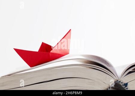 Origami red paper boat floating on the pages of a book on white background. Learning, education, reading, knowledge or conquering adversity concept. Stock Photo