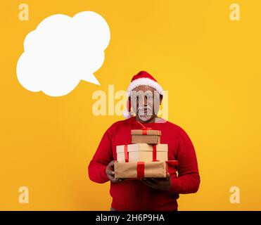 Senior black man wearing Santa hat, holding Xmas gifts, posing with empty speech bubble on orange background, mockup Stock Photo
