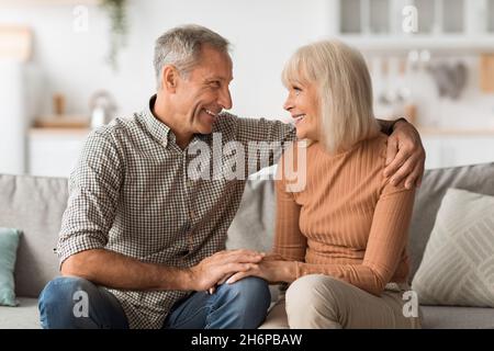 Happy Senior Couple Talking Embracing Sitting On Sofa At Home Stock Photo