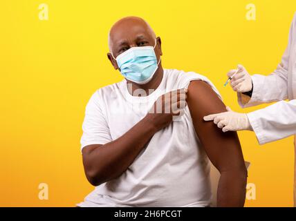 Doctor giving coronavirus vaccine shot to senior African American male patient on orange studio background Stock Photo