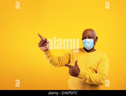 Senior black man in protective face mask pointing at copy space for text or advertisement over orange background Stock Photo