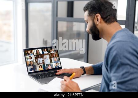 Young Indian businessman man is talking online with group of diverse people, multiethnic team and taking notes with a pen. Multiracial Hispanic male student watching webinar, online classes and writes Stock Photo