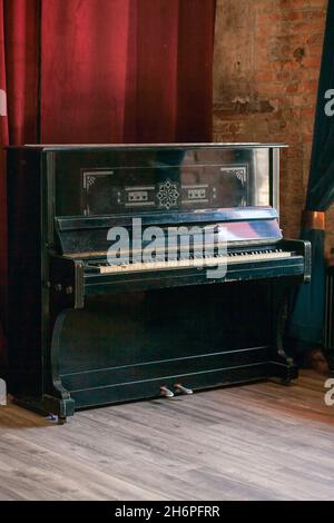 Old vintage piano stands in the hall against the background of a red curtain and a brick wall. Stock Photo