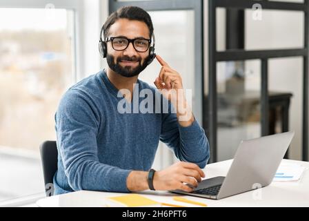 Helpful and smiling Indian male employee wearing headset and eyeglasses making online consultation, holding video call, man in touch looks at camera and touching button on headphones, accepts a call Stock Photo