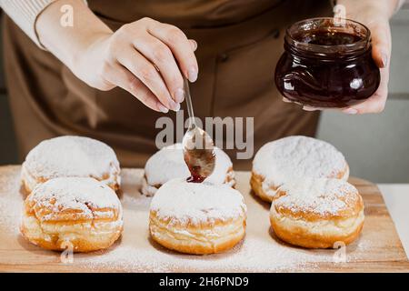 Cooking traditional Hanukkah sufganiyot. A woman decorates donuts with jam. Festive Jewish dessert. Stock Photo