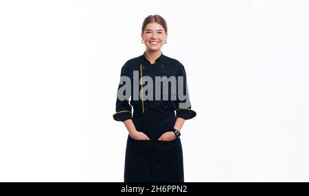 Young woman chef posing at camera, wearing uniform, hands in pockets, smiling