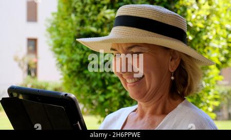 keen Senior female blonde businesswoman in hat uses tablet pc, digital tablet for business work or study in her own green garden. woman aged 50-55 Stock Photo