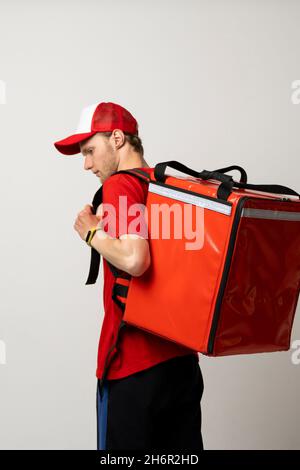 Fast Food Delivery Service Cocnept. Portrait of smiling male courier wearing red uniform holding red thermo backpack bag with a food isolated on white Stock Photo
