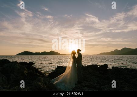 Groom and bride stand on the rocky coast by the sea at sunset Stock Photo