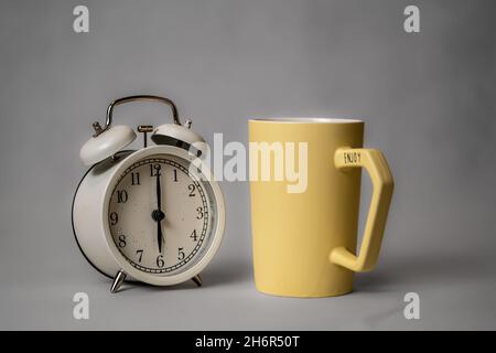 A black alarm clock in retro style and a white coffee cup on a small saucer stand on a pink background. Good morning concept. Morning coffee. Stock Photo