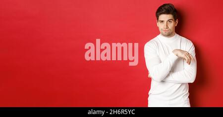 Image of sassy caucasian man in white sweater, looking thoughtful at camera, making choice, standing over red background Stock Photo