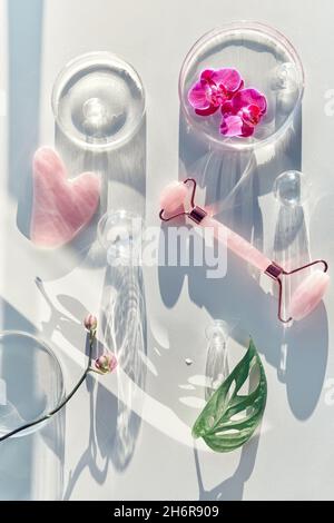 Pink quartz face roller and guasha stone. Face lymph drainage background with exotic monstera leaves, orchid flowers, petri dishes, glass balls Stock Photo