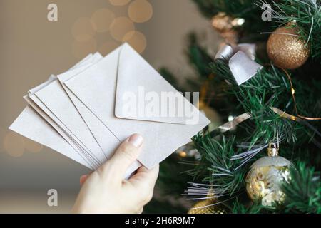 Hand holding stack of craft paper envelopes for christmas greeting card invitation design on christmas tree background with bokeh. Stock Photo