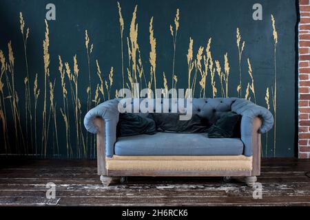 Dark gray quilted old vintage sofa with pillows on a wooden floor near a dark blue wall with painted golden ears Stock Photo