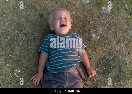 a little boy lies on the ground and cries a lot. resentment or hysteria in a child. Stock Photo