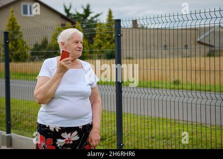 Half length view of an elderly woman talking on a cell phone while standing by the fence Stock Photo