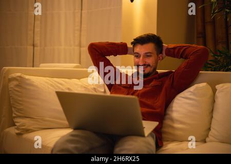 A young man with a beard in a red shirt works with a laptop and sits on the couch in the evening in the house. Stock Photo