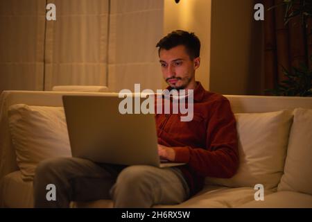 A young man with a beard in a red shirt works with a laptop and sits on the couch in the evening in the house. Stock Photo