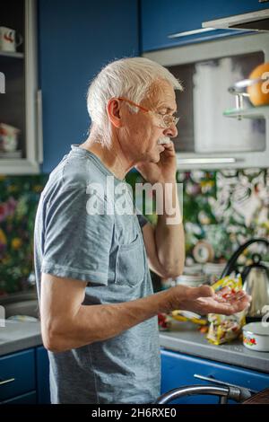 man with gray hair and a mustache speaks on cell phone in the kitchen in everyday life. elderly gray-haired man with a bald head is holding a phone an Stock Photo