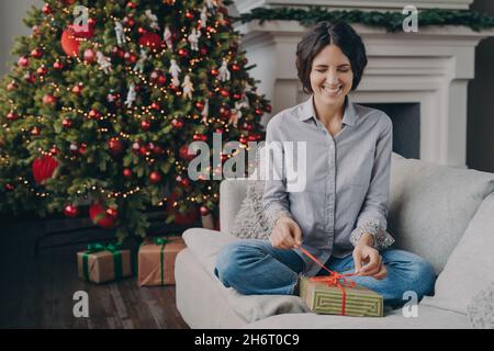 Excited Italian woman with closed eyes trying to unpack Christmas gift while sitting on sofa at home Stock Photo