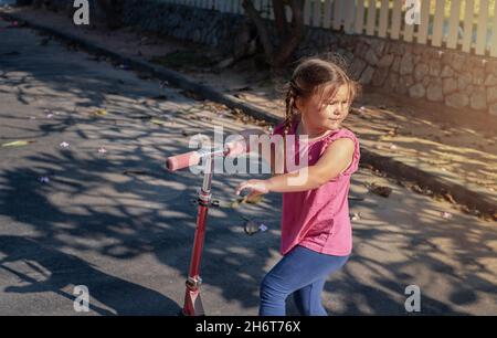 The girl rides a scooter on the road. The child looks back Stock Photo