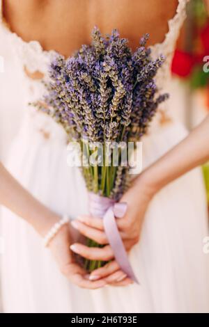 Bride in a white lace dress holds a lavender bouquet tied with a lilac ribbon behind her back. Close-up Stock Photo