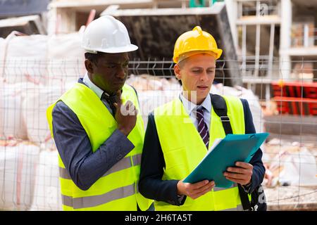 Engineer standing with builder supervising the construction process with house drawings on structure outdoors Stock Photo