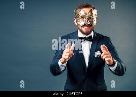 Portrait of man in carnival mask on gray homogeneous background. White Caucasian showman in classic costume looks into camera and smiles emotionally. Face under mask. Stock Photo