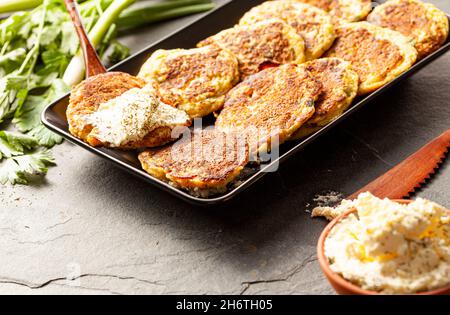 Turkish style zucchini pancakes or fritters known as mucver, served on black plate with fresh greens and sour cream. Close up image on dark stone back Stock Photo