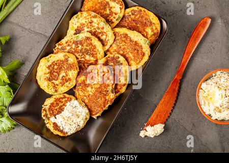 Turkish style zucchini pancakes or fritters known as mucver, served on black plate with fresh greens and sour cream. Close up image on dark stone back Stock Photo