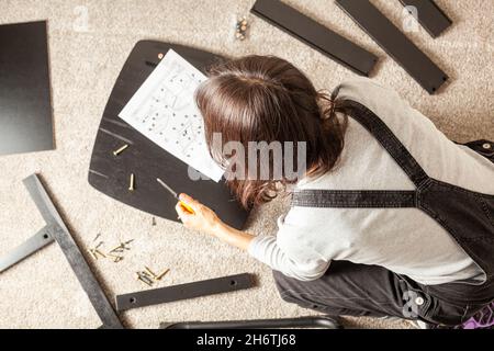 DIY furniture assembly concept including a caucasian person sitting on carpet floor with pieces of furniture, trying to assemble it based on instructi Stock Photo