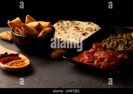 Close up image of a table with delicious dishes from India. Low light dark background with naan, curry chicken, palak paneer, samosa, various spices a Stock Photo