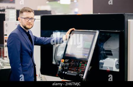 Engineer young man in glasses operates CNC automatic metalworking lathe or laser, industrial worker in modern factory. Stock Photo