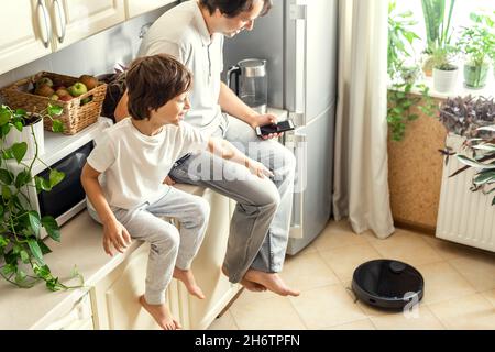Happy Man and his Son sitting on the kitchen table top testing the Robotic Vacuum Cleaner for the first time at Home. Remote Controling with Smartphon Stock Photo