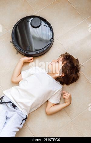 Kid playing with Robotic Vacuum Cleaner. Boy lying on the floor in the way of the Vacuum Cleaner route Stock Photo