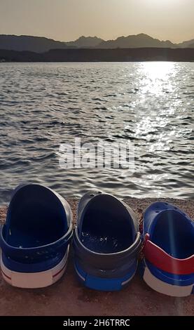 Three multi-colored sandals of different shapes lie in a row on the beach by the sea. Stock Photo