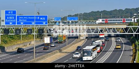 Greater Anglia passenger train crossing railway bridge above road traffic junction 28 M25 motorway for A12 Brentwood Essex & gantry signs England UK Stock Photo