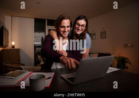 Caucasian happy couple working from home typing on laptop with arms around each other  Stock Photo
