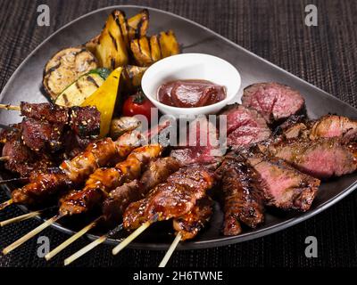 meat plate, set of different grilled skewers and grilled sliced filet mignon on a plate served with grilled vegetables, close up photo on a dark backg Stock Photo