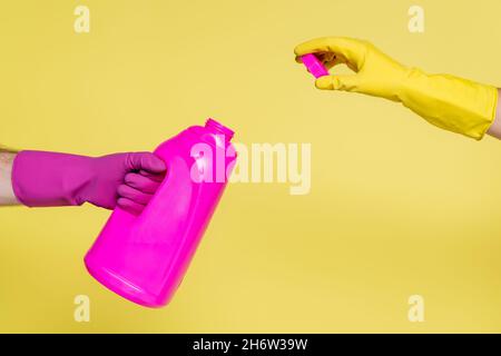 cropped view of people in rubber gloves holding pink bottle with detergent and bottle cap isolated on yellow Stock Photo