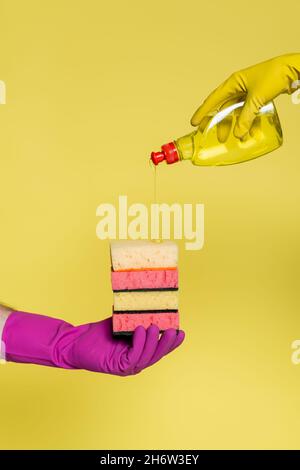 cropped view of people in rubber gloves holding bottle with detergent and stack of sponges isolated on yellow Stock Photo